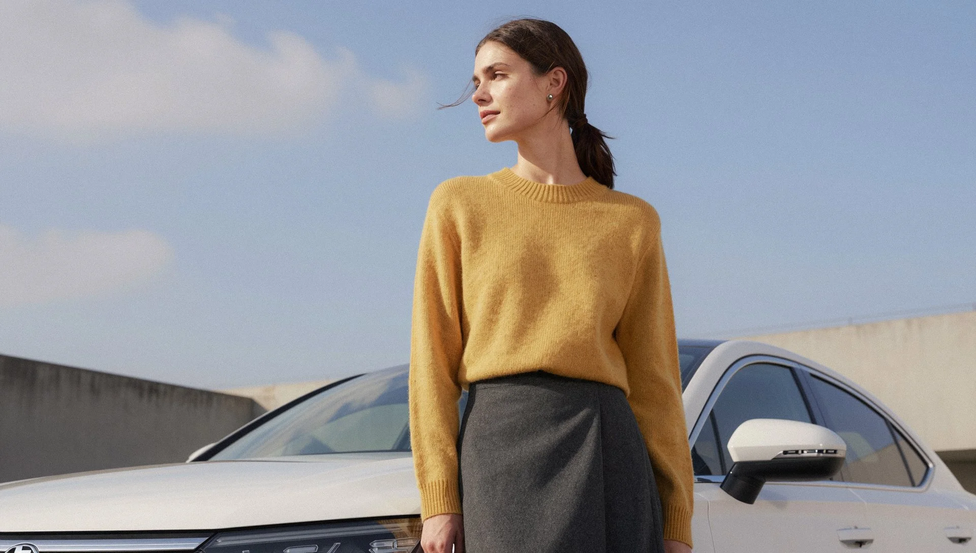 Woman in a yellow sweater and gray skirt stands outdoors beside a white car on a partly cloudy day.