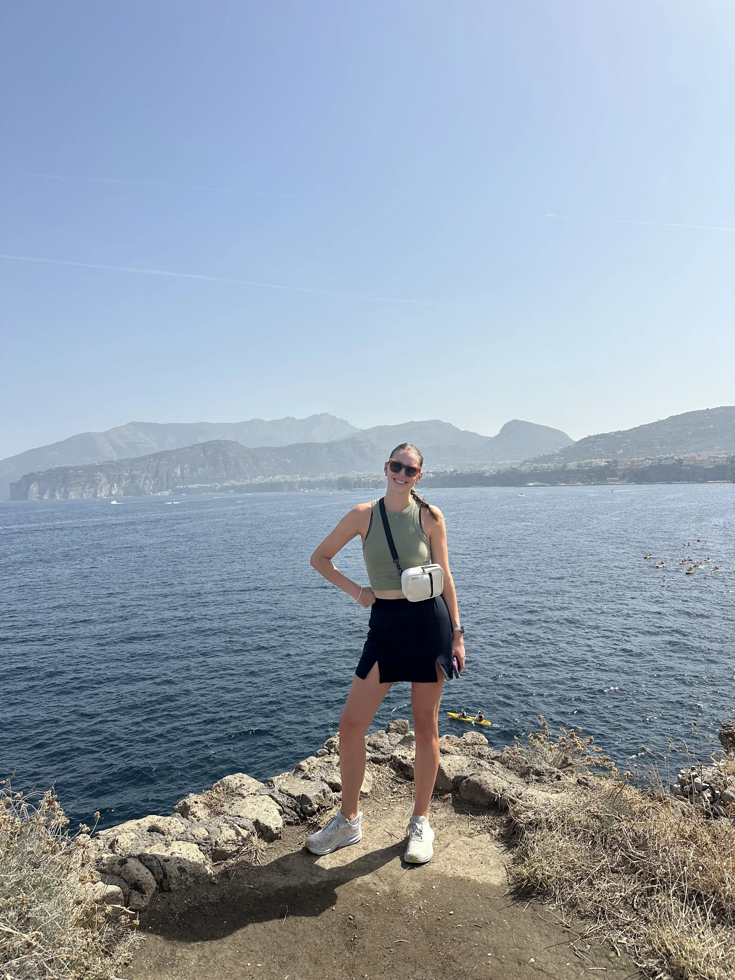 A woman standing on rocky ground near water with mountains in the background, wearing sunglasses, a sleeveless top, a skirt, and sneakers, smiling at the camera.