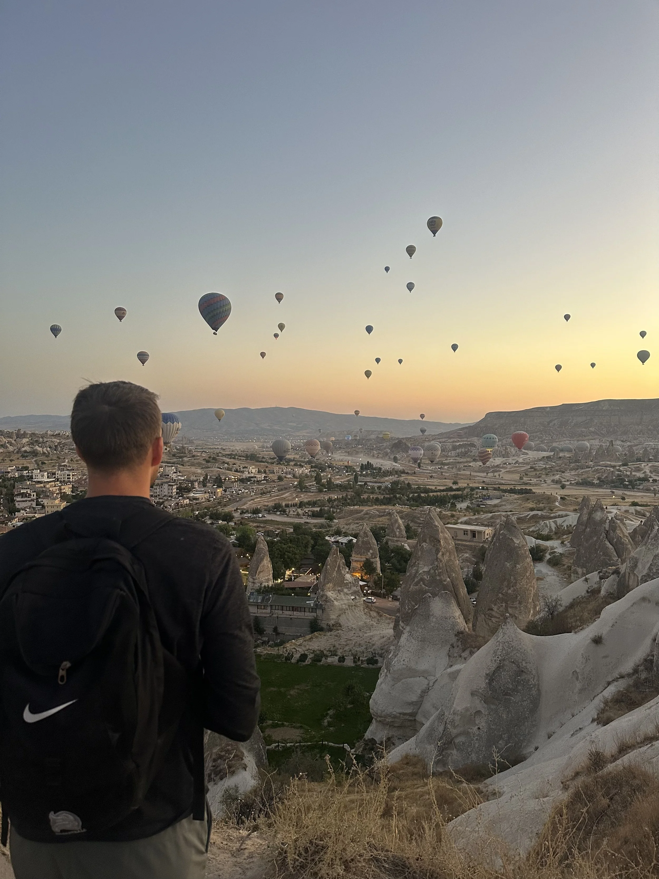 A man with a black Nike hoodie and a backpack stands on rocky terrain overlooking a landscape with numerous hot air balloons in the sky during sunset.