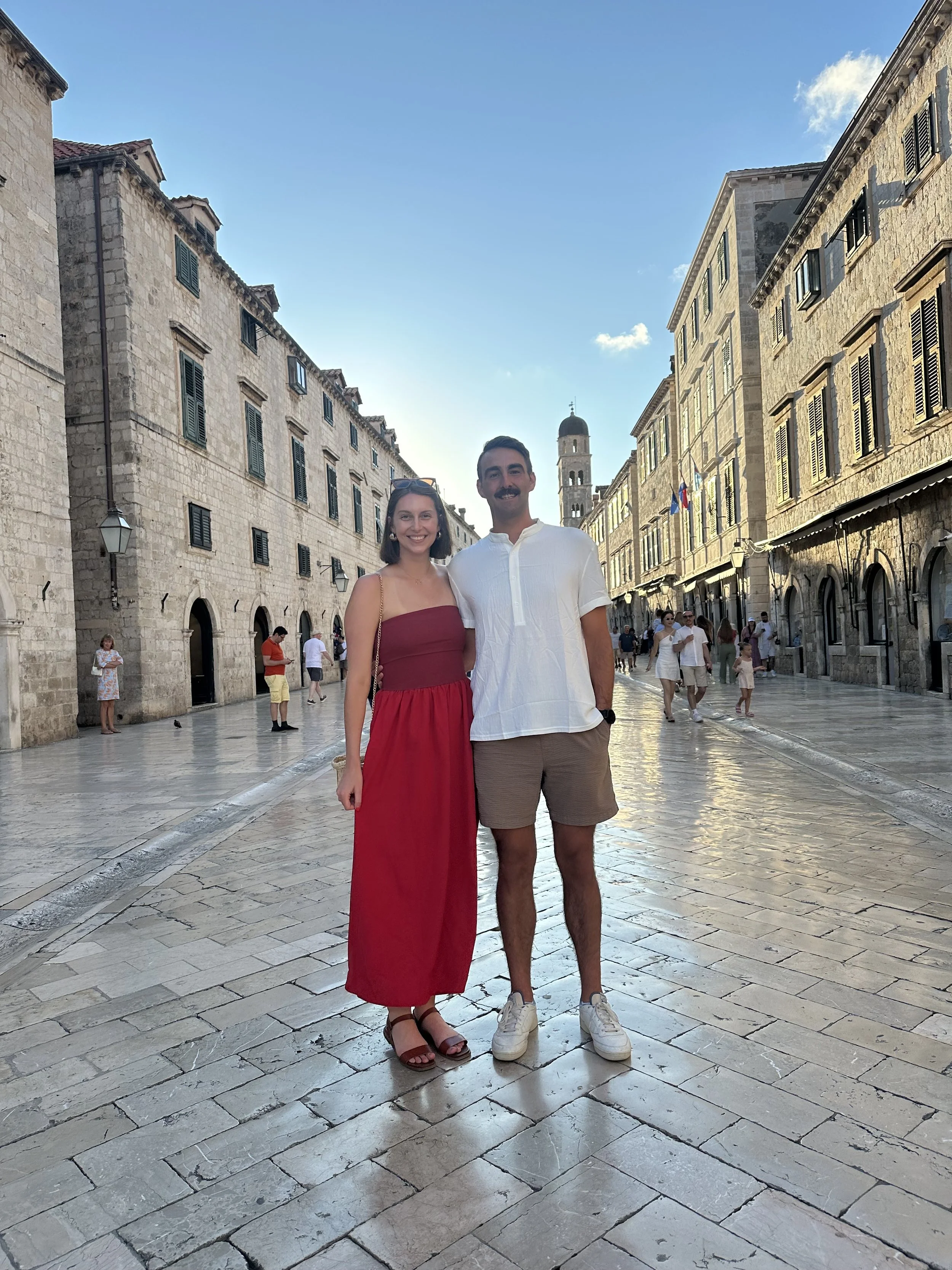 A smiling woman in a red dress and a smiling man in a white shirt and khaki shorts standing on a cobblestone street in an old European city, with historic stone buildings and a tower in the background.