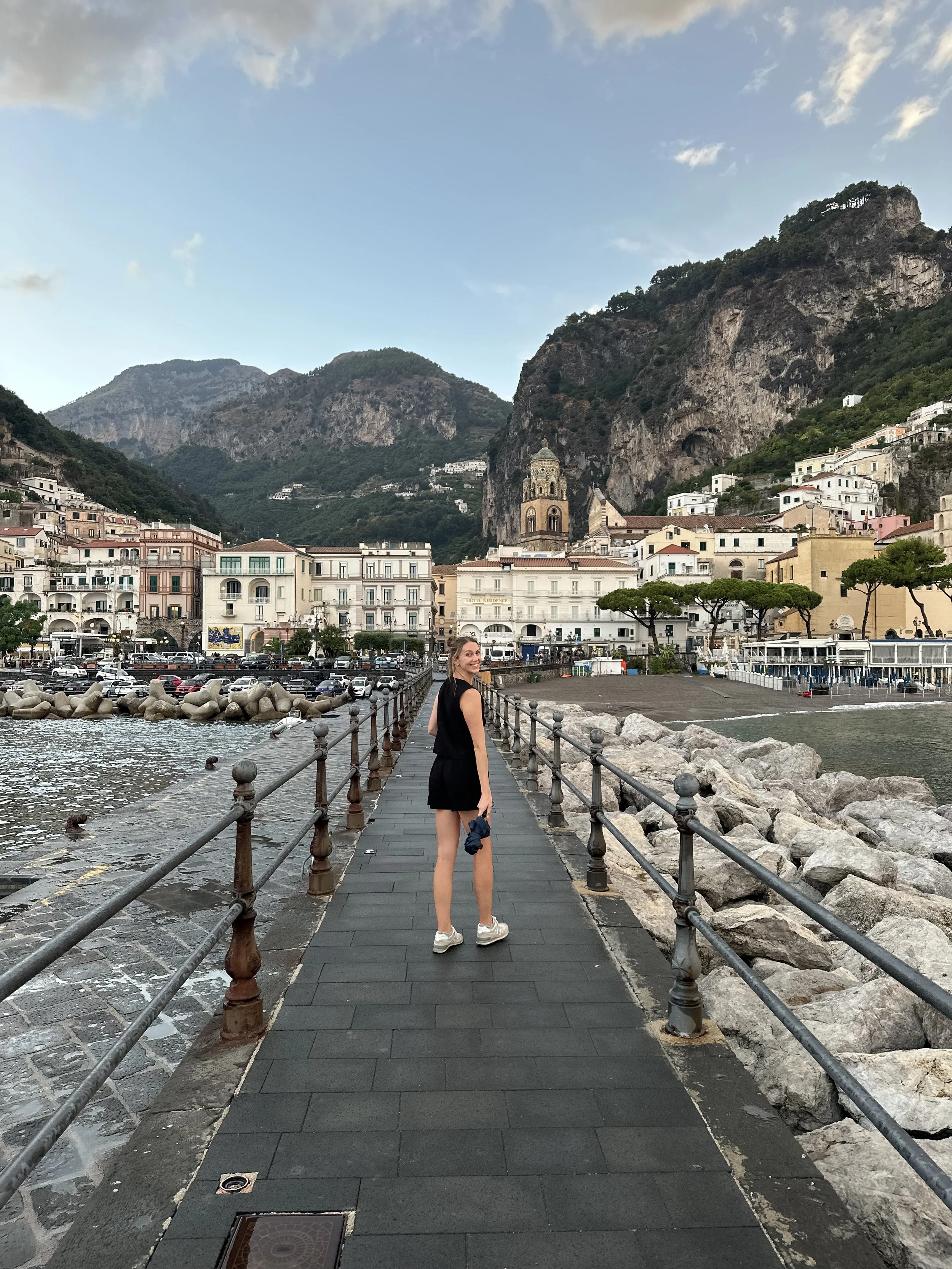 A woman with light brown hair wearing a black sleeveless dress and beige sneakers standing on a waterfront promenade, looking back and smiling. Behind her are multi-story buildings, a church tower, and a steep hillside with greenery and white houses. The scene is set against a mountainous landscape with a partly cloudy sky.