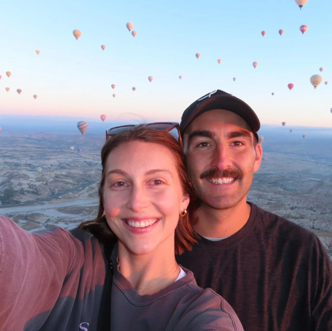 A smiling woman and man taking a selfie during a hot air balloon ride at sunset, with numerous balloons floating in the sky behind them and a landscape below.