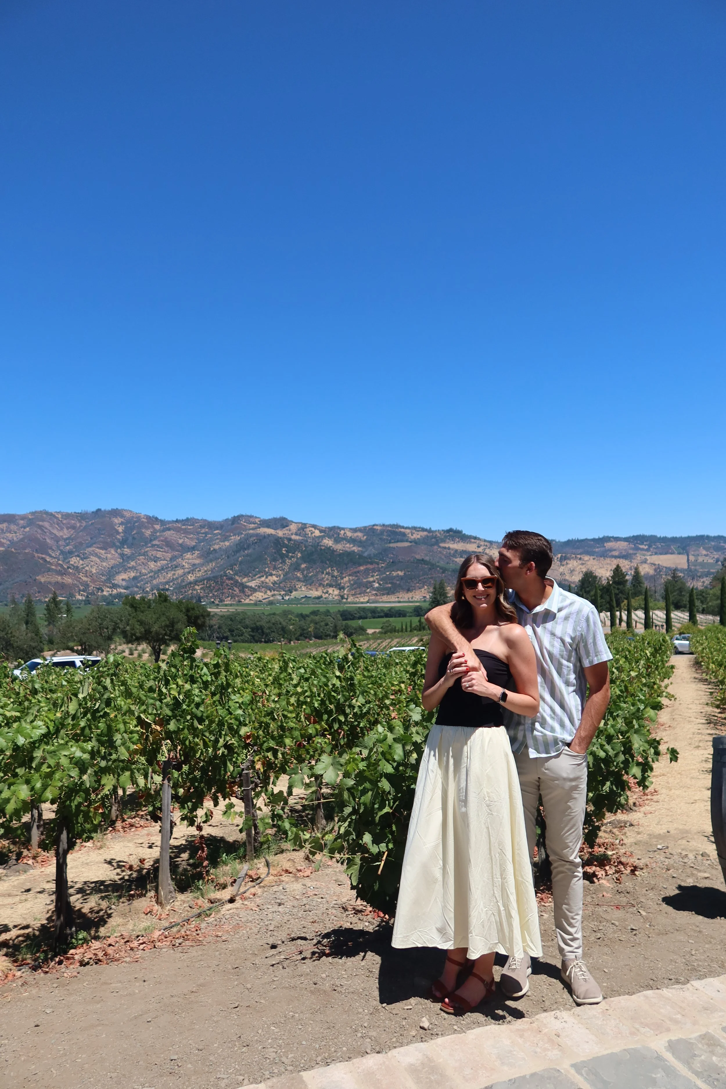 A smiling woman in a black strapless top and cream-colored skirt stands next to a man in a striped shirt and light pants, in a vineyard with green grape vines, mountains, and a clear blue sky in the background.