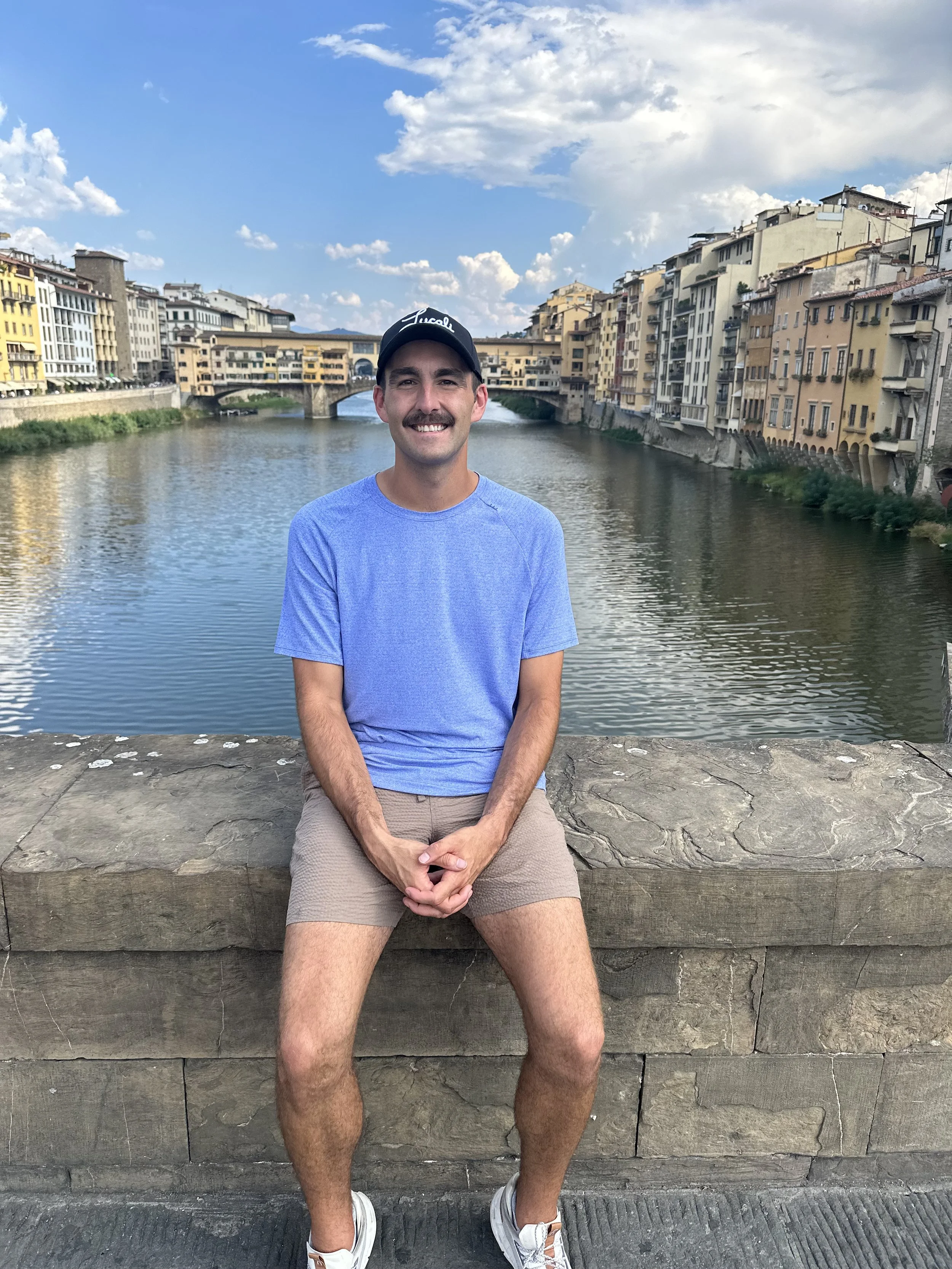 A man with a mustache sitting on a stone wall by a river in a city with historical buildings and a bridge, under a blue sky with clouds.