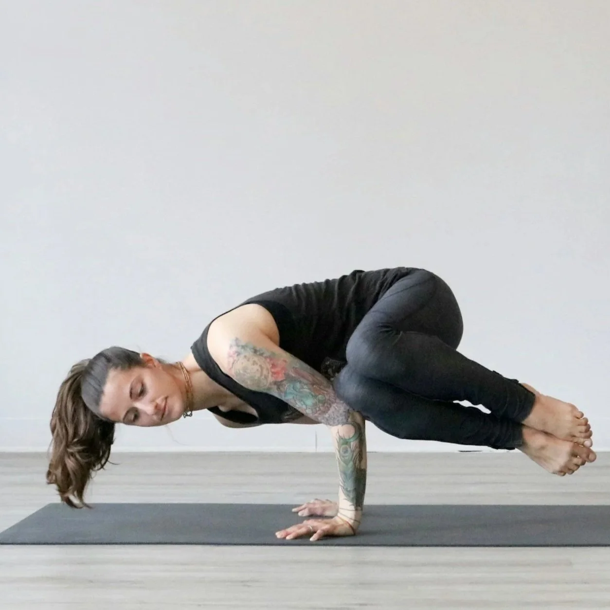 Woman performing a yoga pose on a black mat, balancing on her hands with knees tucked towards her chest, in a room with light-colored walls.