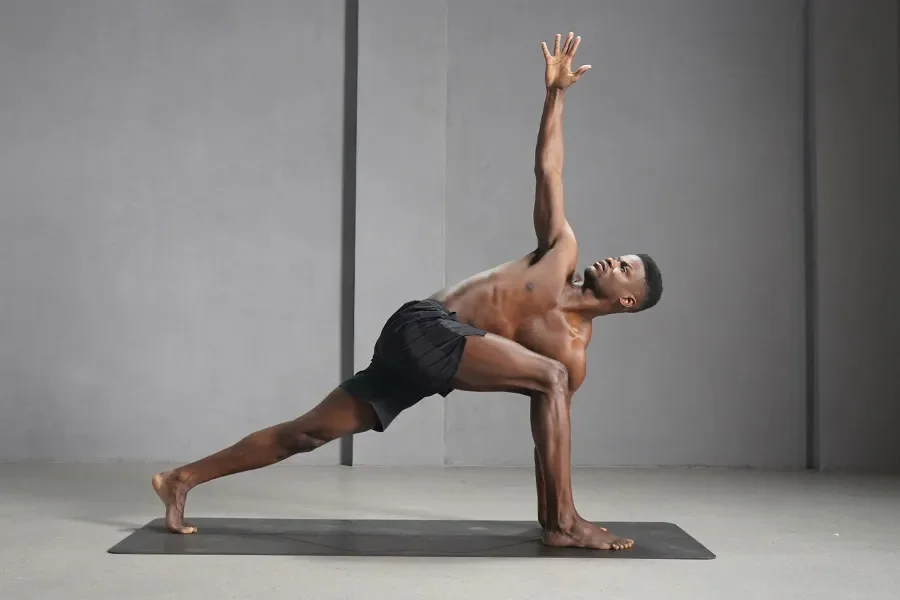 A practitioner moves through a grounded, strength-based pose — one leg extended, one arm reaching upward — in a calm, minimal space designed for focused practice.