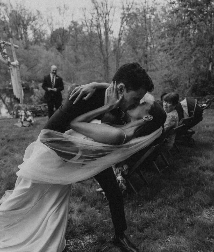 A bride and groom share a passionate kiss outdoors during a wedding ceremony, with a man in the background and a woman sitting on a bench nearby.