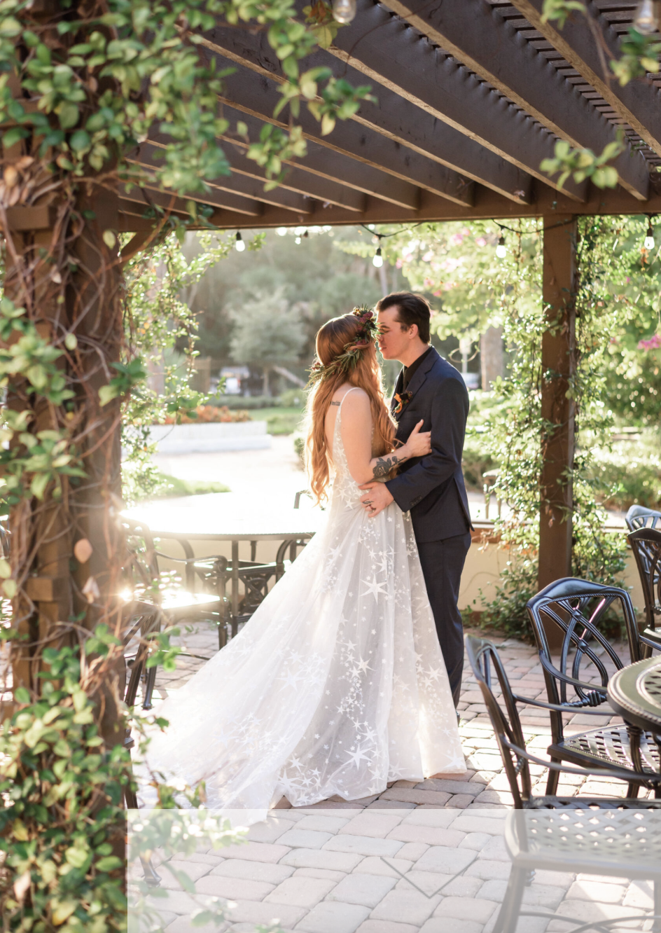 A bride and groom share an intimate moment under a wooden pergola decorated with greenery and string lights outdoors. The bride has long red hair, a floral crown, and a white gown with star patterns. The groom has dark hair and is wearing a dark suit. Sunlight filters through the scene, creating a romantic setting.