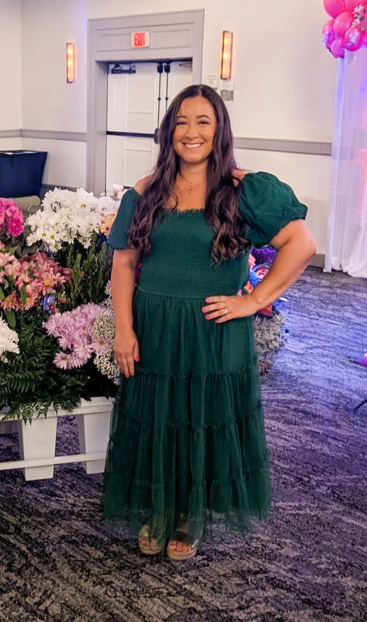 A woman smiling and standing with one hand on her hip in a room decorated with flowers, pink balloons, and sheer pink and white curtains.