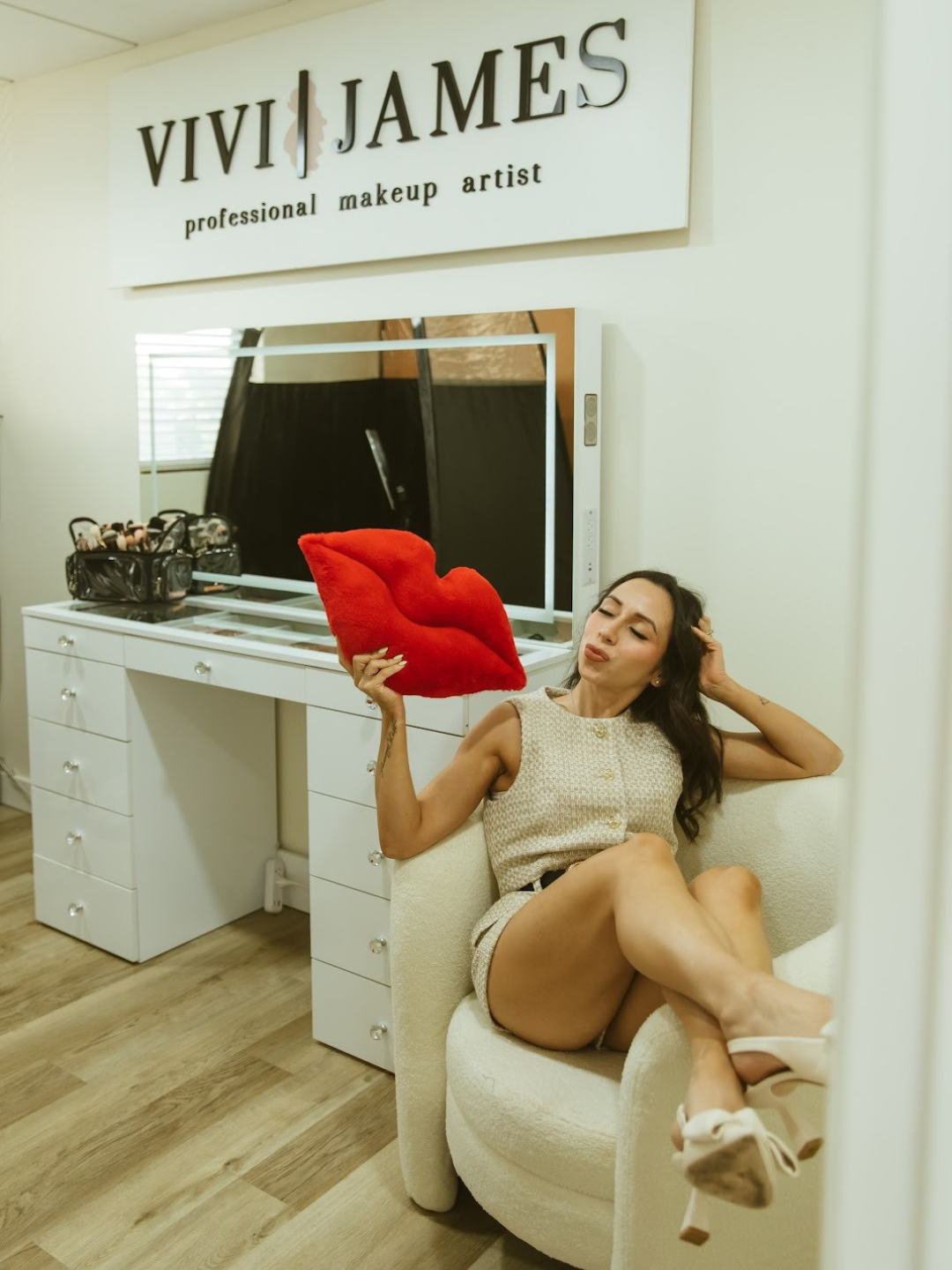 A woman sitting on a white chair, holding a red pillow shaped like lips, in a makeup studio with a sign reading 'VIVI JAMES professional makeup artist.'