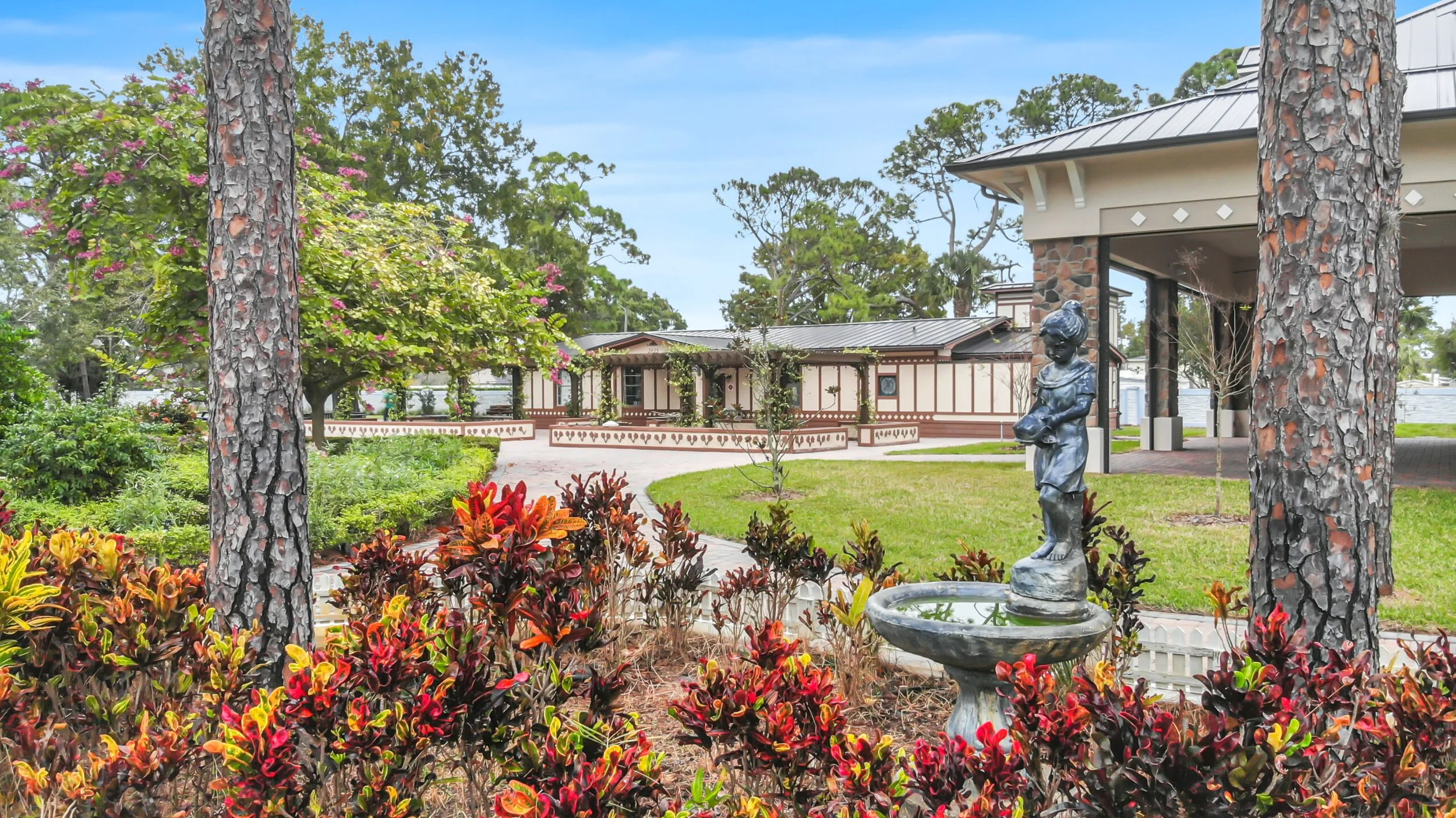 A landscaped garden with colorful shrubs, tall pine trees, a flowering tree with pink blossoms, a stone fountain with a statue of a girl, a neatly maintained lawn, and a building with a metal roof and wooden trim in the background.