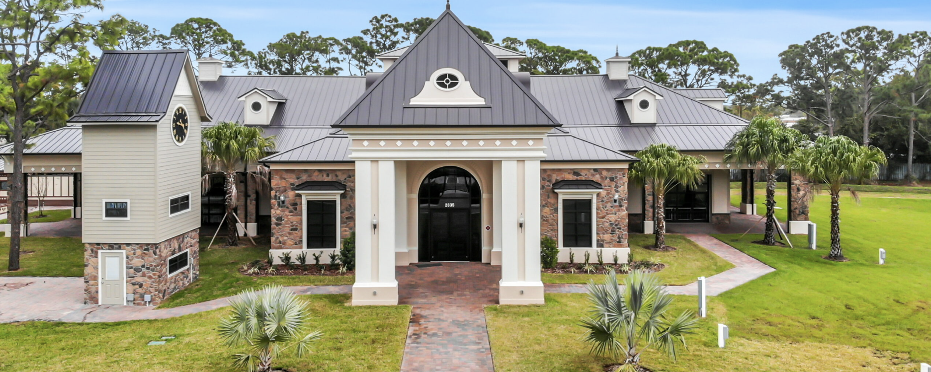 A large modern house with a stone and siding exterior, multiple gables, and a pitched metal roof, surrounded by a well-maintained lawn with palm trees, brick pathways, and a cloudy sky.