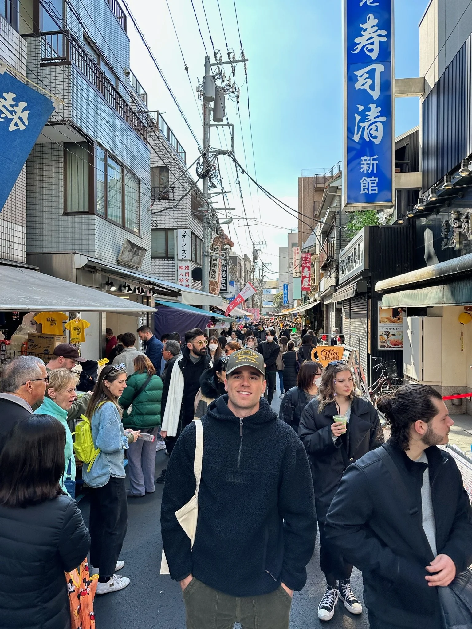 Crowded street market with various shops, signs in Japanese, and people walking and shopping. A young man in a black hoodie and cap is smiling in the foreground.