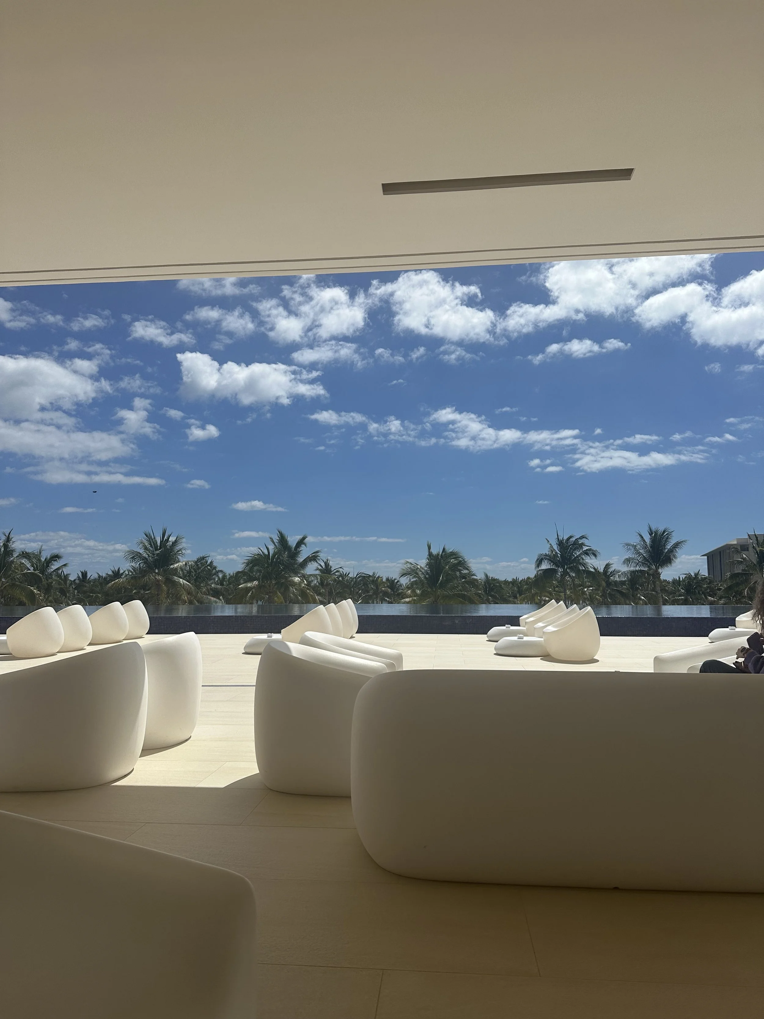 Outdoor seating area with white modern chairs and palm trees in the background under a blue sky with clouds.