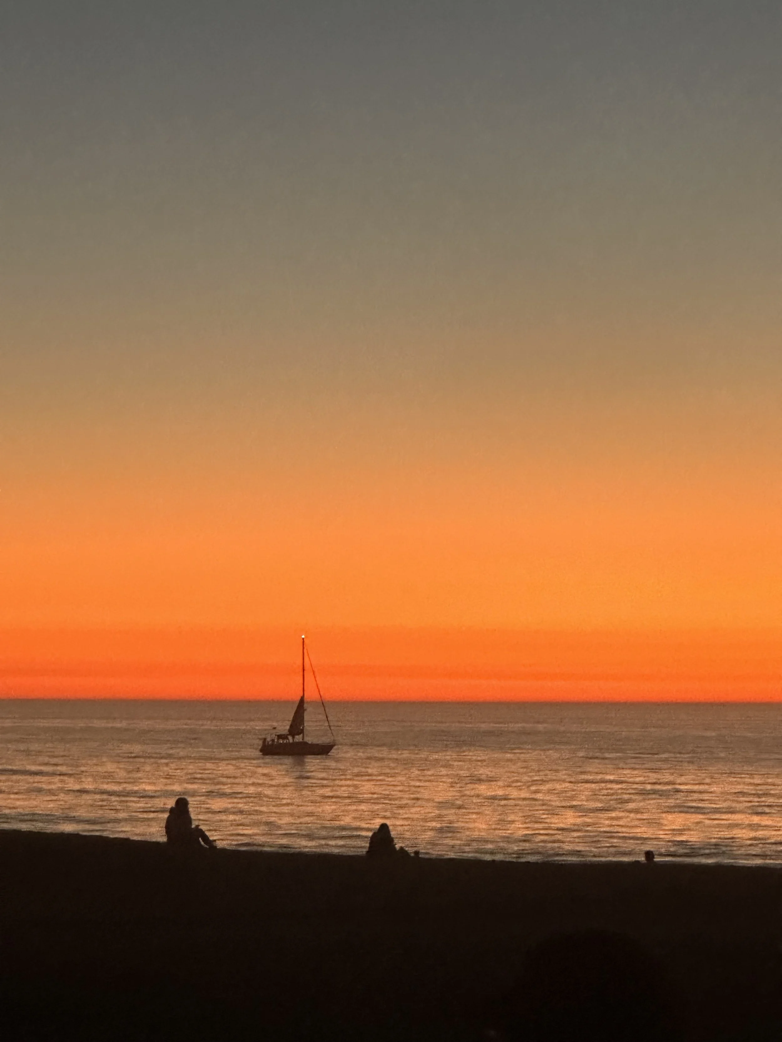 A sunset over the ocean with an orange and blue sky. A sailboat is on the water and people are sitting on the beach in silhouette.
