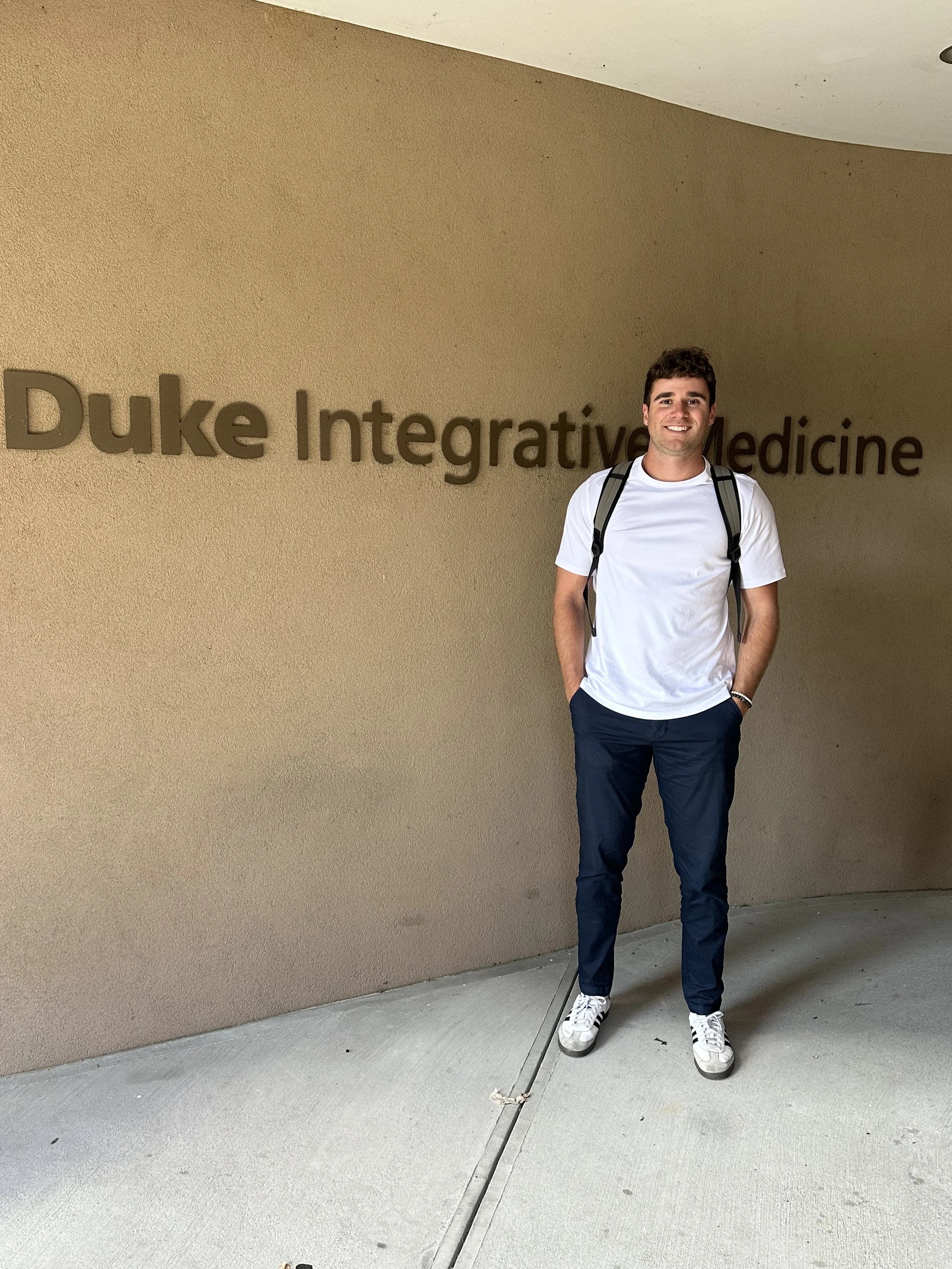 A young man standing in front of a beige wall with the sign 'Duke Integrative Medicine,' smiling, wearing a white T-shirt, navy blue pants, white sneakers, and carrying a backpack.