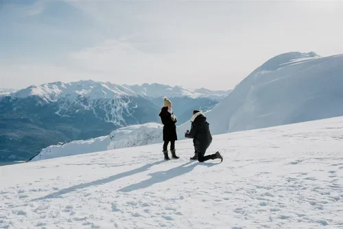 Two people standing on snow-covered ground in a mountainous, snowy landscape