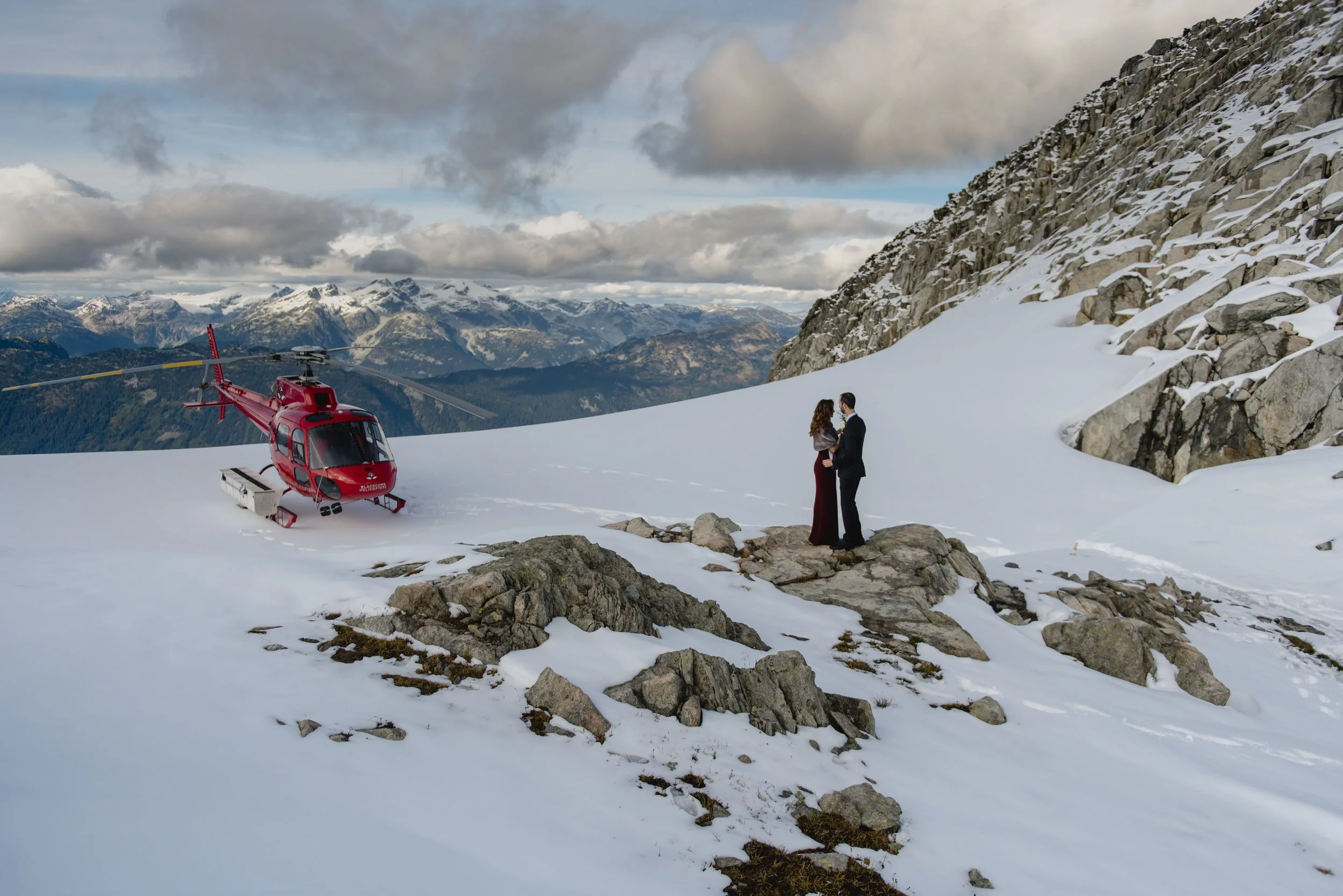 Couple standing on mountain summit next to red helicopter during heli-wedding.
