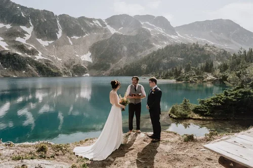 A wedding ceremony taking place by a mountain lake with two people in wedding attire and an officiant