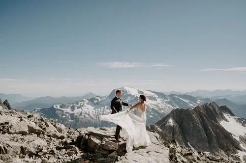 A bride and groom holding hands on a mountain peak with snow-capped mountains in the background.