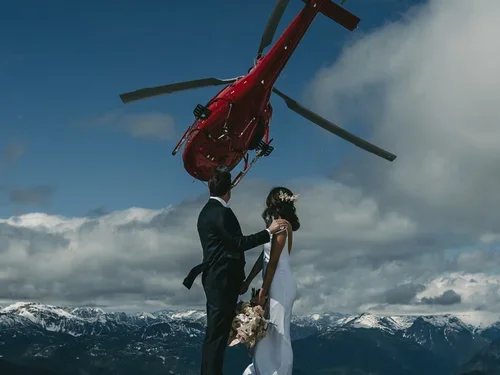 A bride and groom holding hands outdoors with snow-covered mountains in the background, a red helicopter flying overhead, and cloudy sky.