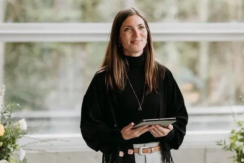 A woman with long brown hair, wearing a black turtleneck and black jacket, holding a tablet, standing in front of a large window with greenery outside.