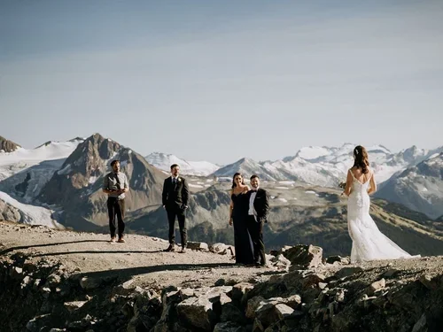 People dressed in formal attire standing on rocky terrain in a mountain landscape with snow-covered peaks.
