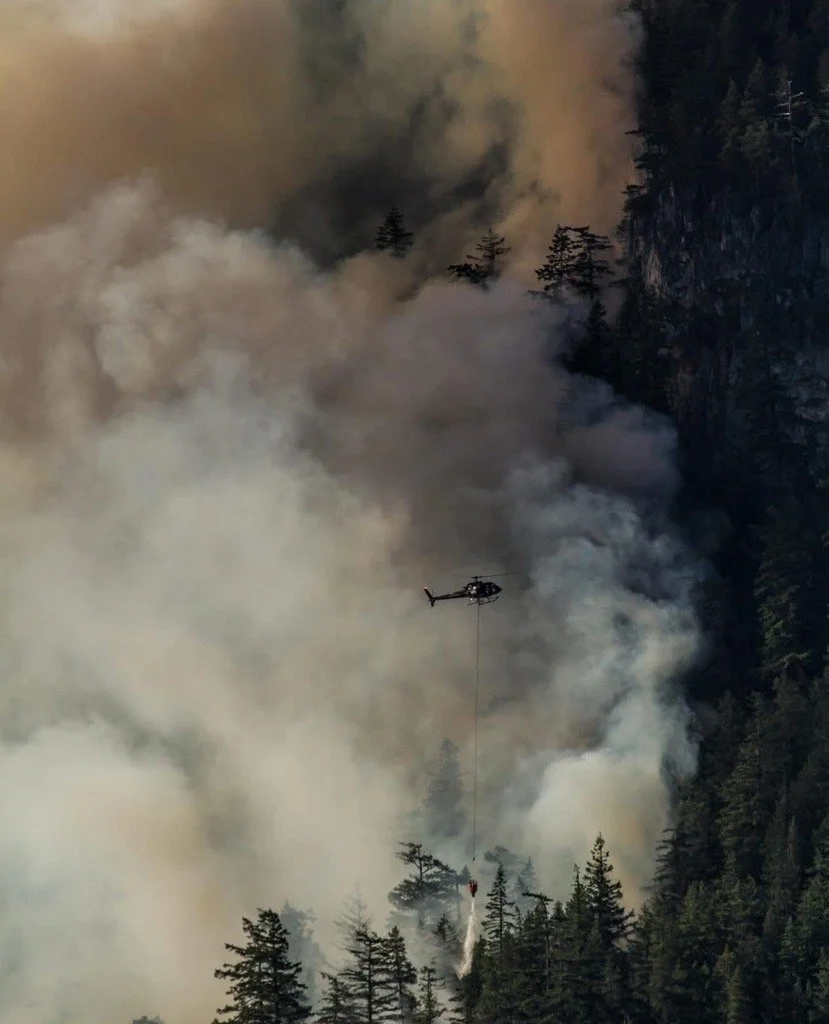 Helicopter dropping water over wildfire surrounded by smoke.
