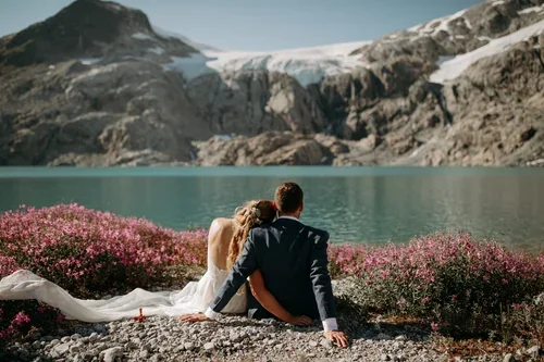 Couple sitting by a lake with mountains and a glacier in the background, surrounded by pink flowers.