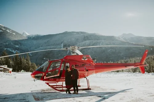 Two people standing near a red helicopter on snowy ground with mountains in the background.
