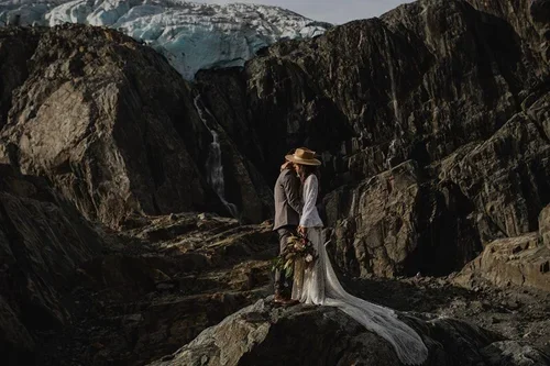 A couple in wedding attire standing on rocky terrain near a glacier, surrounded by large dark rocks.