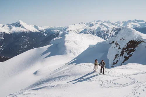 Two people hiking on a snow-covered mountain ridge with snowy peaks in the background.