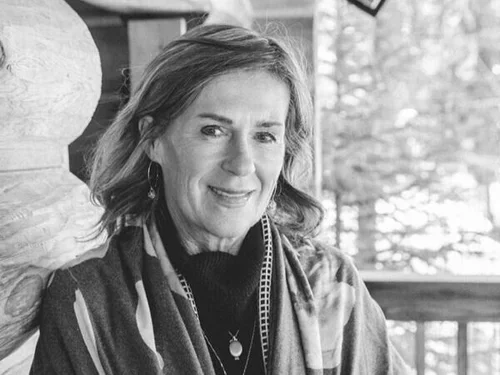 A woman with shoulder-length hair smiling, wearing a scarf and earrings, sitting indoors with a window and trees visible in the background.