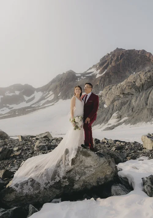 A bride and groom standing on a large rock in a snowy mountainous landscape during their wedding photoshoot. The bride is wearing a white wedding gown holding a bouquet, and the groom is dressed in a burgundy suit. The mountains in the background are snow-capped with rugged terrain.