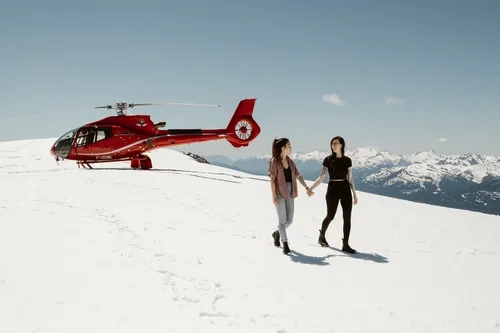 Two women holding hands walking on snow with a red helicopter and snow-capped mountains in the background.