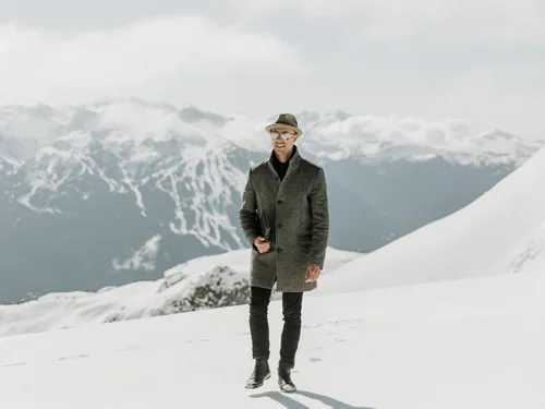 A person standing on snowy mountainous terrain with snow-capped peaks in the background, dressed in a dark coat, black pants, and a hat.
