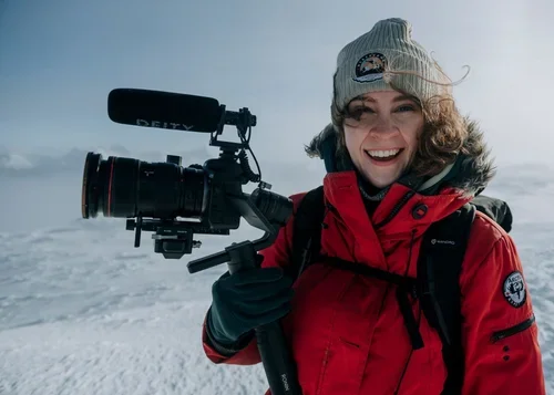 Woman in red winter jacket and gray knit hat holding professional video camera outdoors on snowy landscape.