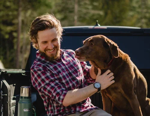 A man with a beard and plaid shirt sitting on a truck bed, smiling and holding a brown dog with a red collar in an outdoor setting.