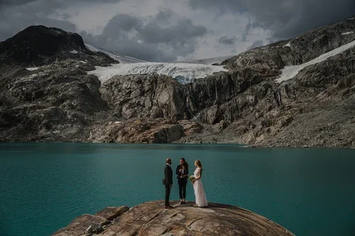 A wedding ceremony taking place on a rocky area by a large alpine lake with mountains and glaciers in the background under cloudy skies.