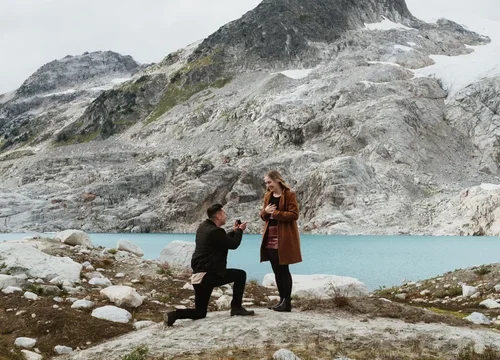 A man proposing marriage to a woman in a scenic mountain landscape with a lake and rocky terrain.