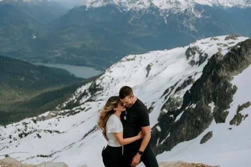 A couple hugging in front of a snowy mountain landscape with a lake and mountain peaks in the background.
