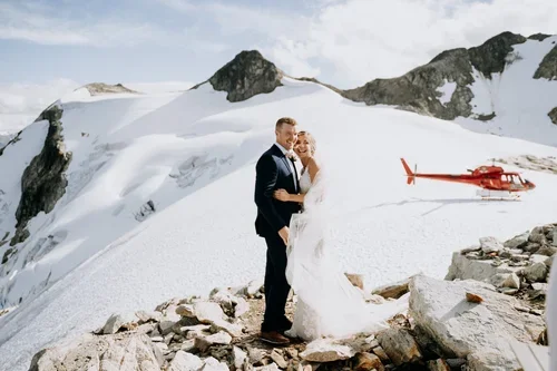 A newlywed couple standing on snowy mountain terrain with a red helicopter in the background under cloudy sky.