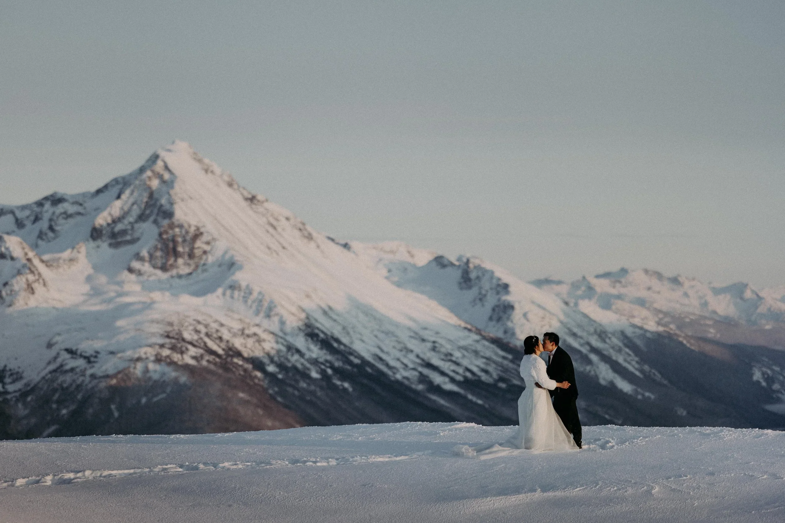 Bride and groom kissing on snowy mountain during heli-wedding.