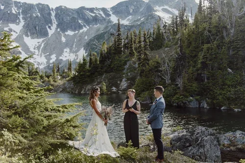 A wedding ceremony taking place outdoors by a river in a mountainous forest setting, with a bride in a white gown, a groom in a dark suit, and a female officiant.