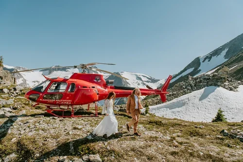 A red helicopter on a rocky mountain landscape with snow-covered peaks in the background, a woman in a white dress, and a man in a brown suit.