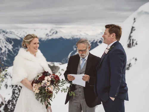 A bride and groom standing outdoors in a snowy mountain landscape during their wedding ceremony, with an officiant reading from a paper.