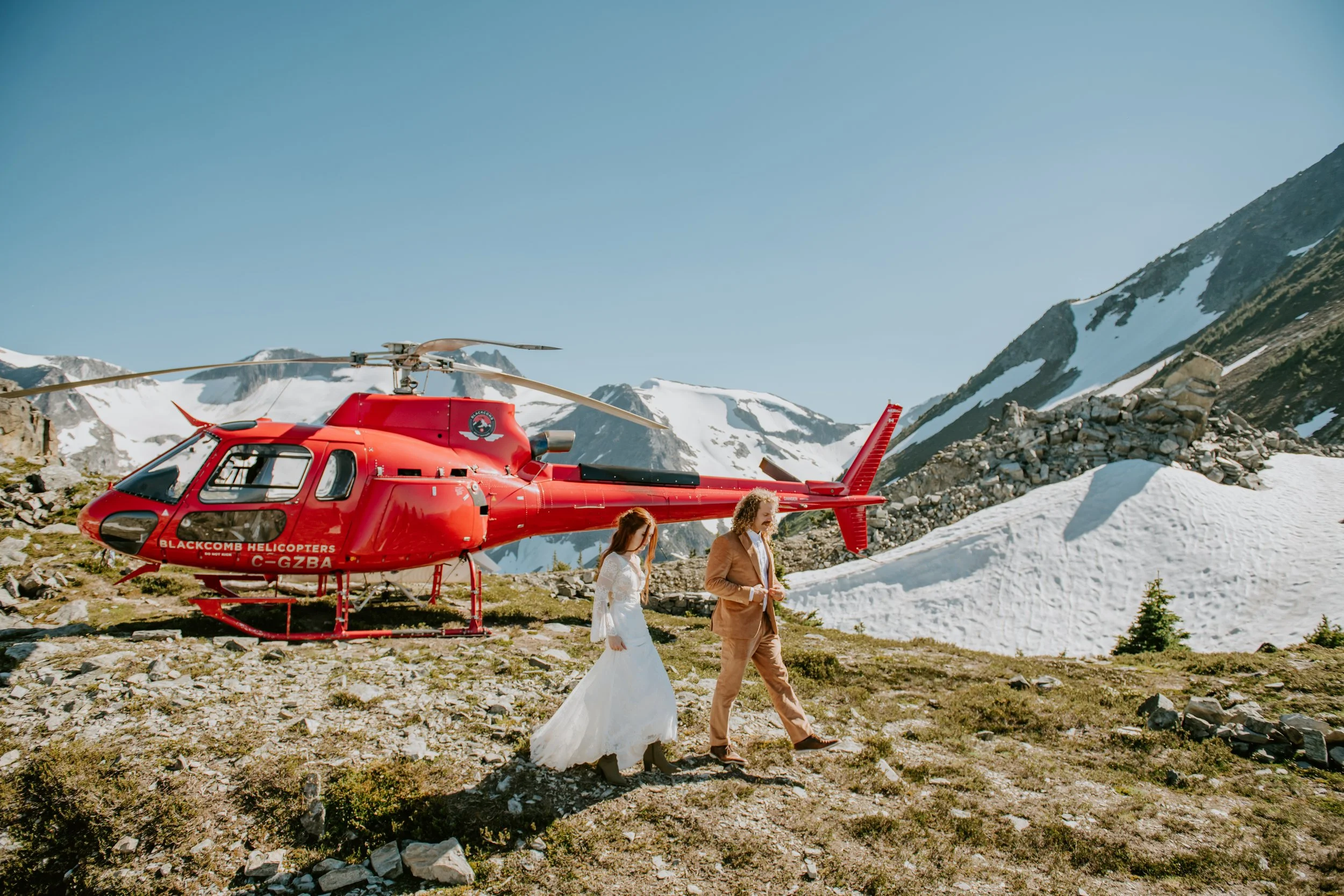 Bride and groom walking near red helicopter with summer mountains in background.