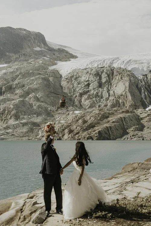 A bride and groom holding hands near a mountain lake, with a helicopter flying over rocky mountains in the background.