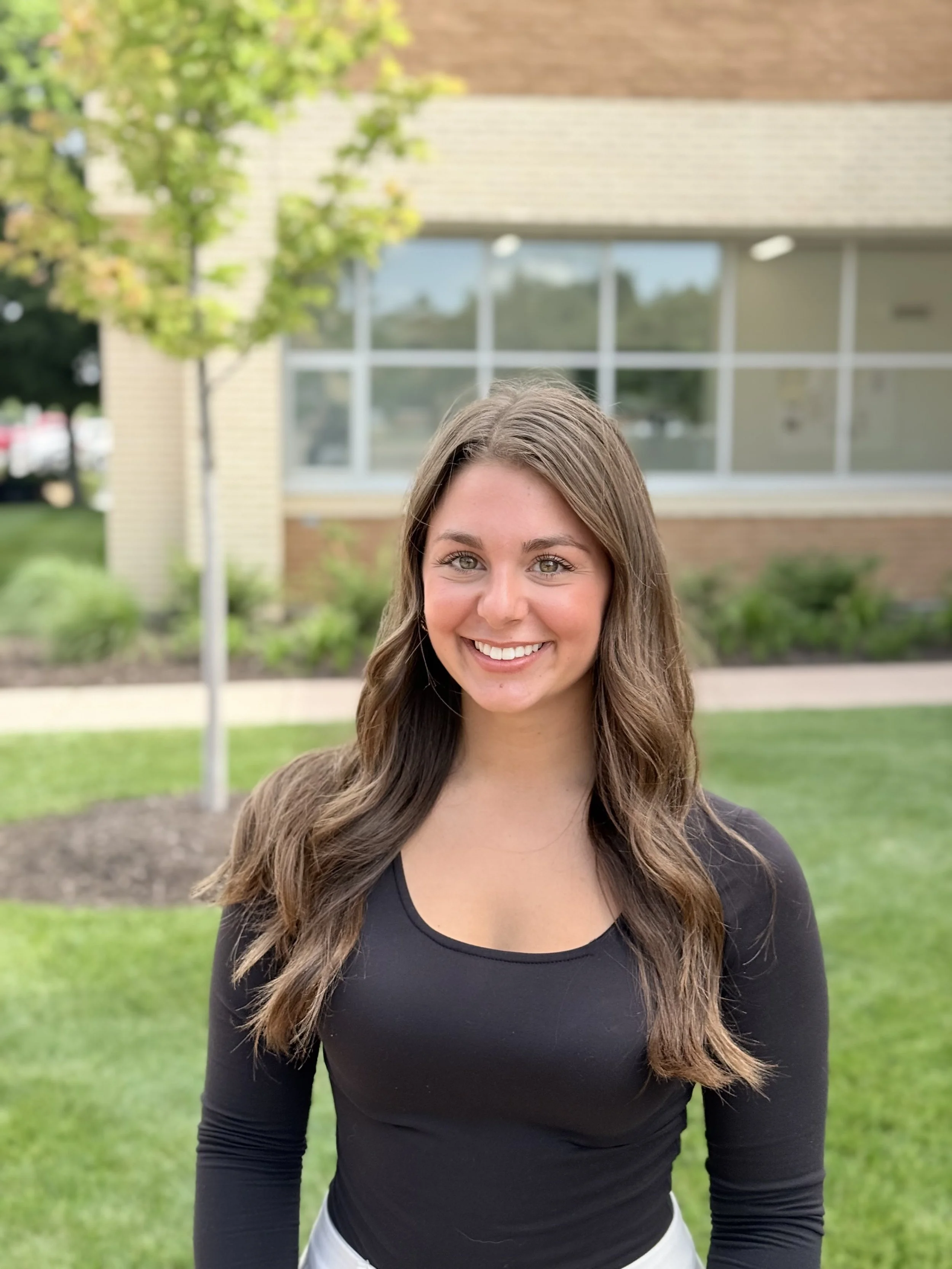 A young woman with wavy brown hair, wearing a black top, smiling outdoors in front of a building with large windows and a small tree.