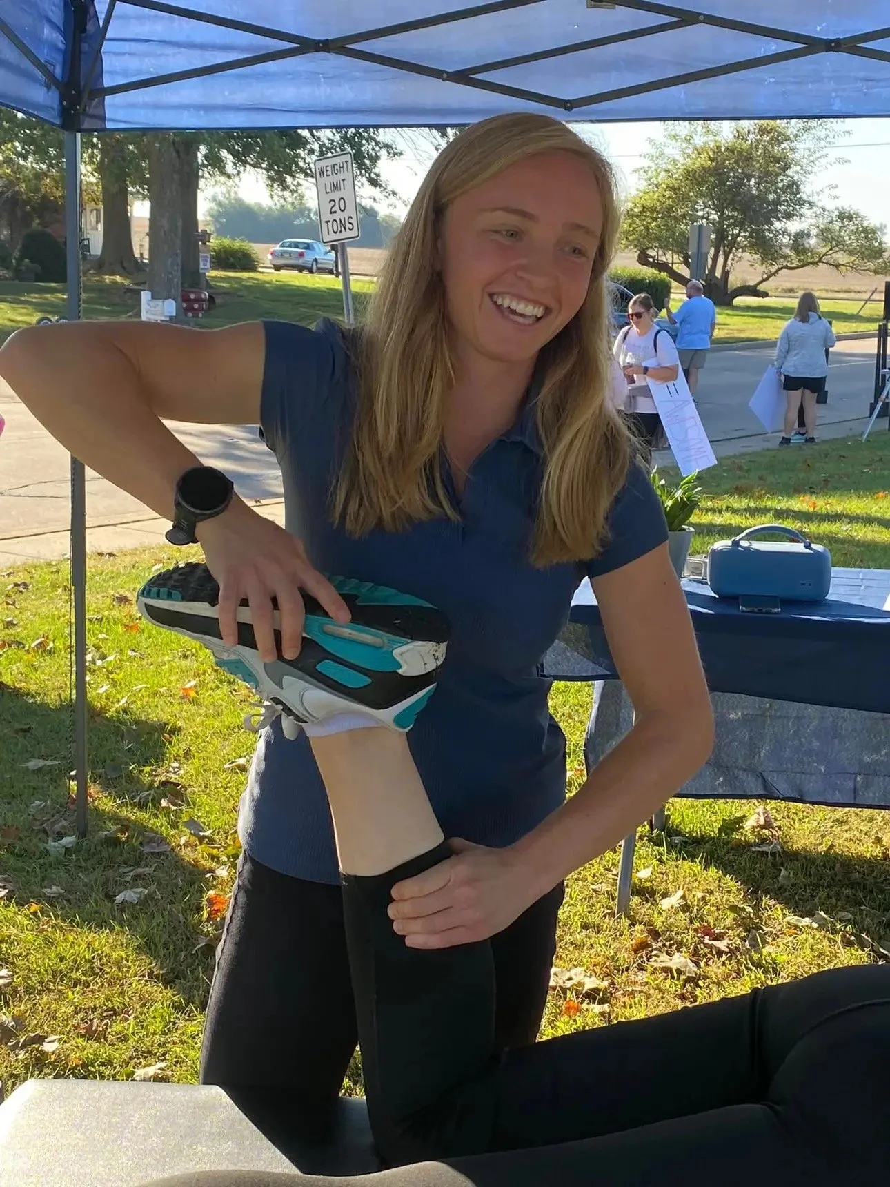 A woman is smiling and stretching her leg, which is resting on a table, outdoors under a blue tent. She is wearing a dark blue polo shirt and black pants. Other people are visible in the background at an outdoor event.